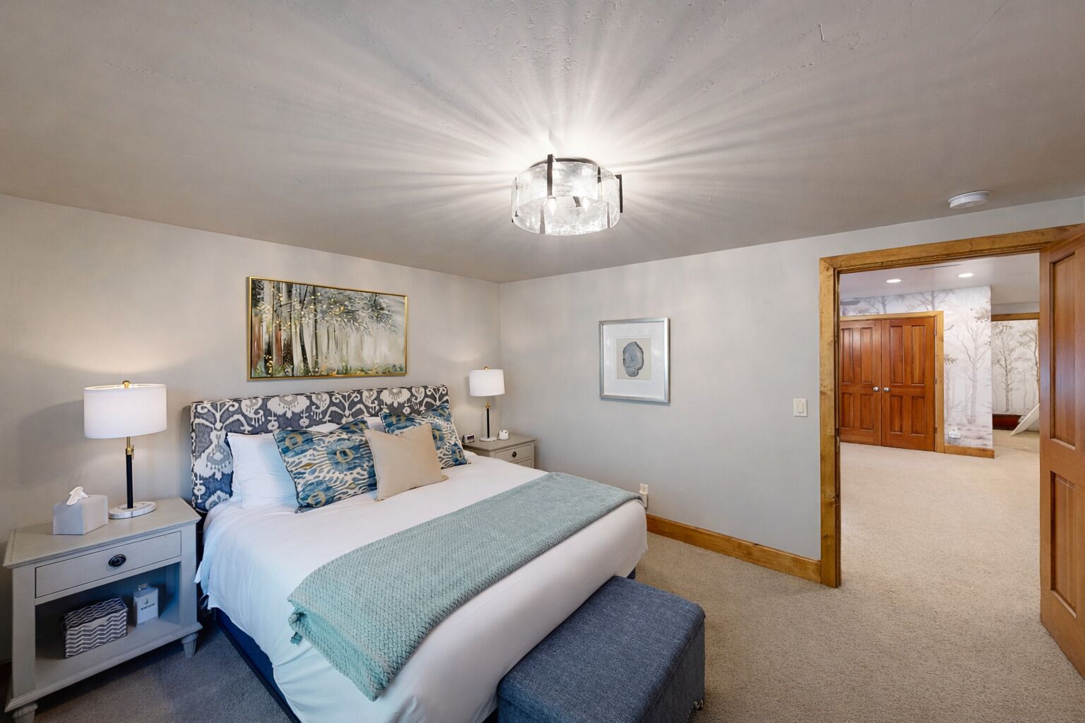 Un dormitorio con cama, mesitas de noche, una lámpara, un cuadro, una fotografía enmarcada y una puerta que da a otra habitación.