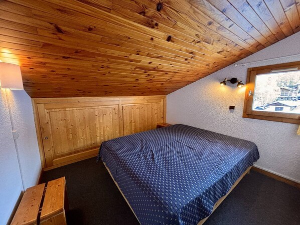 Een kleine, knusse kamer met een schuine kap, houten wanden, een enkel bed, een bankje en een raam met uitzicht.