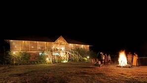 A large building with a staircase leading to a balcony, surrounded by a grassy area and a campfire. - Sixth Element (Somvarpet)