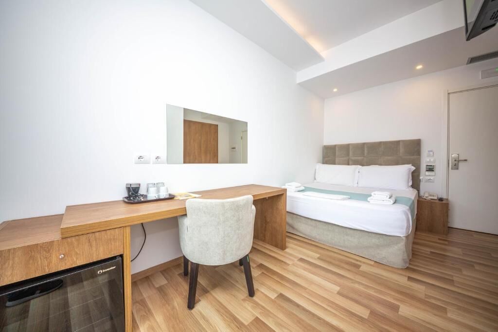 A modern hotel room with a wooden desk, a bed with white linens, a small bedside table, and a built-in oven.