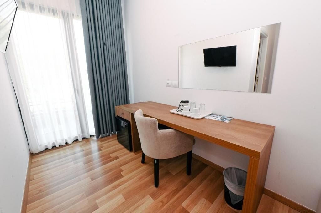 A hotel room with a wooden desk, a chair, a small trash bin, a mirror, and a television mounted on the wall.