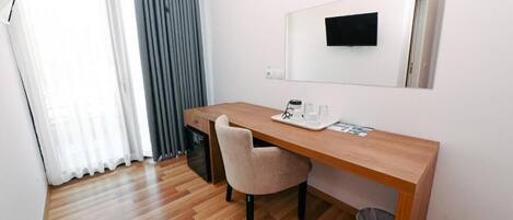 A hotel room with a wooden desk, a chair, a small trash bin, a mirror, and a television mounted on the wall.