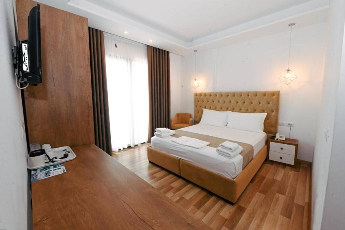 A hotel room with a large bed, wooden flooring, a TV mounted on the wall, and a bedside table with a lamp.