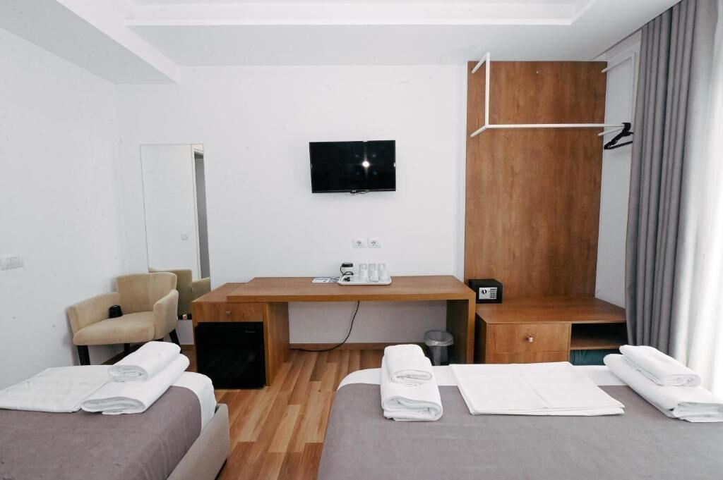 Ein modernes Hotelzimmer mit Schreibtisch aus Holz, einem an der Wand befestigten Fernseher, einem Sessel und zwei Betten mit Handtüchern sowie einem kleinen Mülleimer.