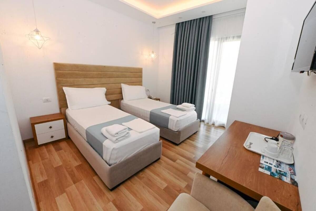 A hotel room with two beds, a wooden headboard, a small bedside table, a wooden desk, and a window with curtains.