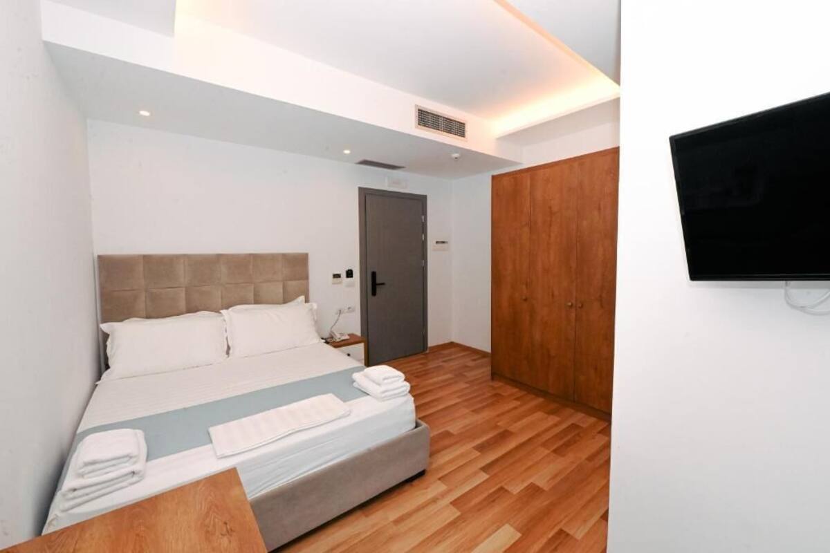 A modern hotel room with a large bed, a wooden wardrobe, a TV mounted on the wall, and a small bedside table.