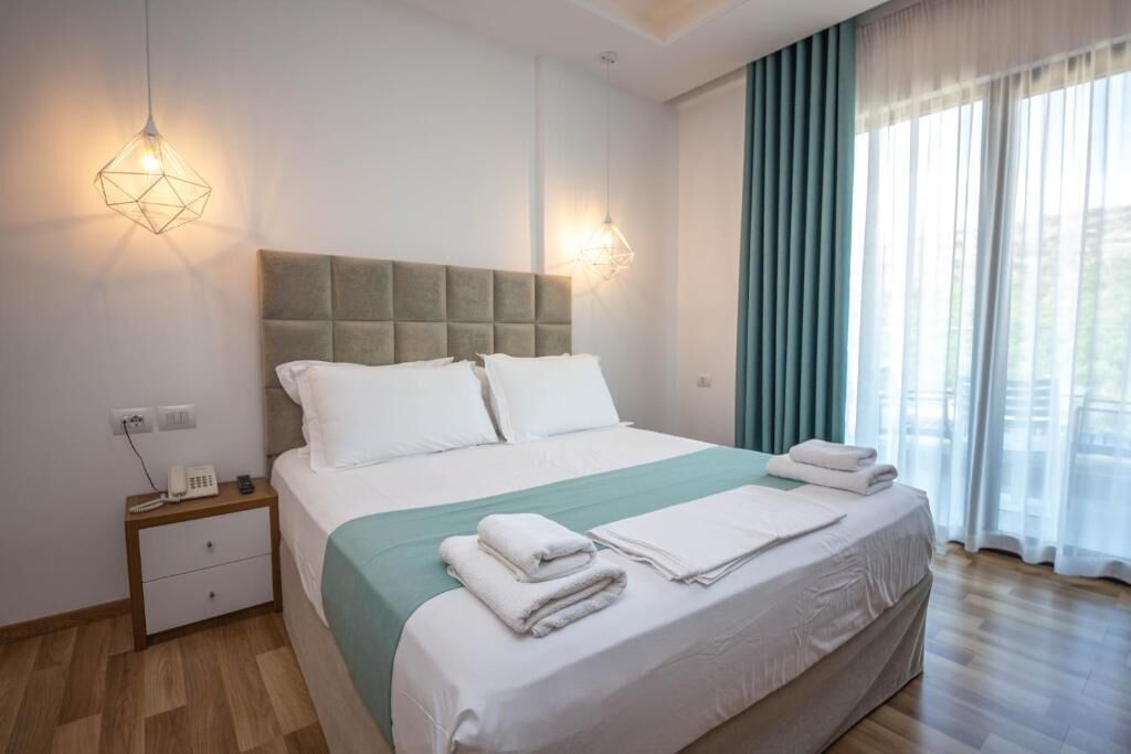 A neatly made bed with white linens and a teal bedspread, a bedside table with a phone, and a window with sheer curtains.