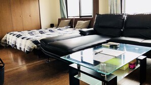 A modern living room with a sofa, a glass coffee table, a bed with a checkered duvet, and a wooden wardrobe.