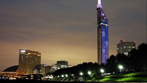 A high-rise building with illuminated windows and a distinctive spire, surrounded by other buildings and a park area with street lamps.