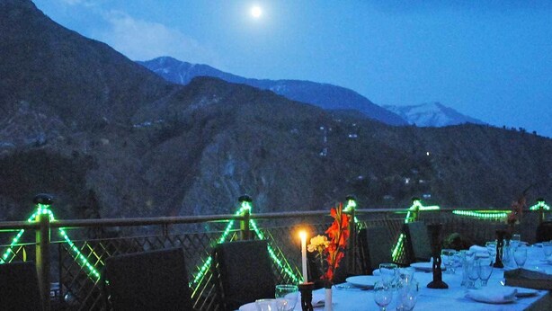 Comedor con mesas dispuestas para recibir a los comensales, con vistas a un paisaje montañoso bajo un cielo nocturno y una luna visible.