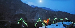 Comedor con mesas dispuestas para recibir a los comensales, con vistas a un paisaje montañoso bajo un cielo nocturno y una luna visible.