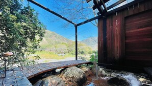 Una estructura de madera con un puente de piedra sobre un pequeño arroyo, rodeada de árboles y colinas.