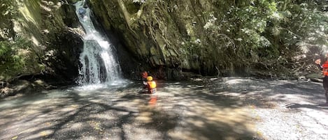 A waterfall in a forested area with a person in a yellow helmet and orange vest standing near the water.
