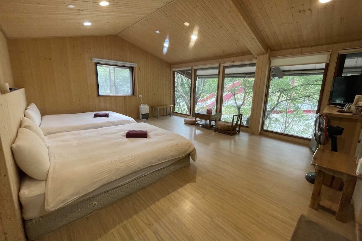 A spacious room with two beds, large windows, a wooden ceiling, and a television on a wooden stand.