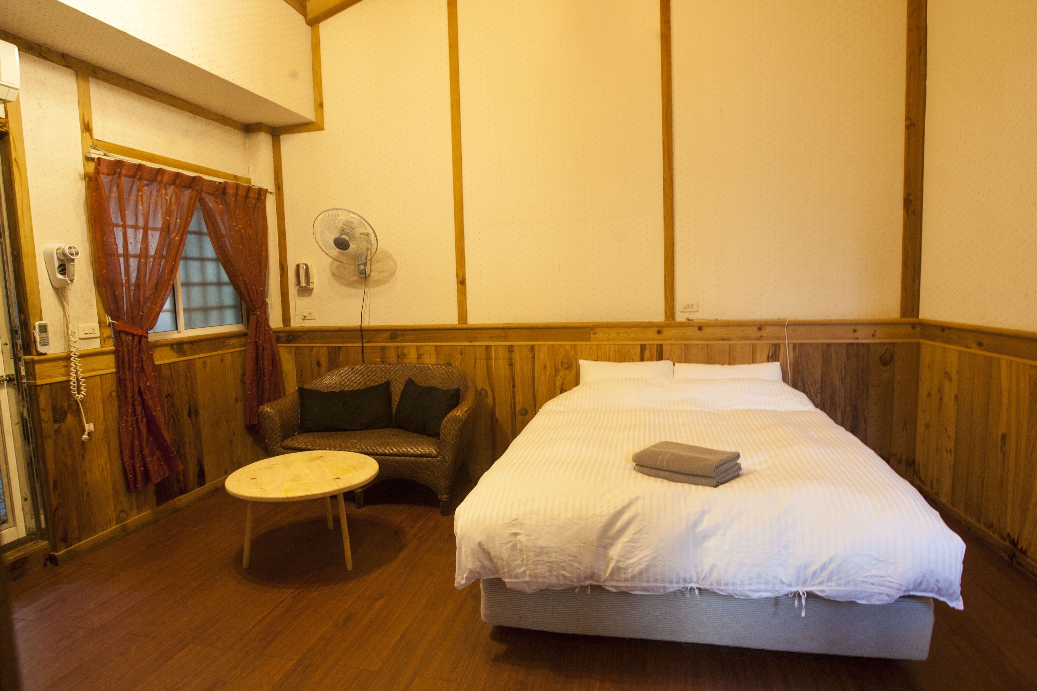 A room with a bed, a chair, a small table, a fan, and a window with curtains.