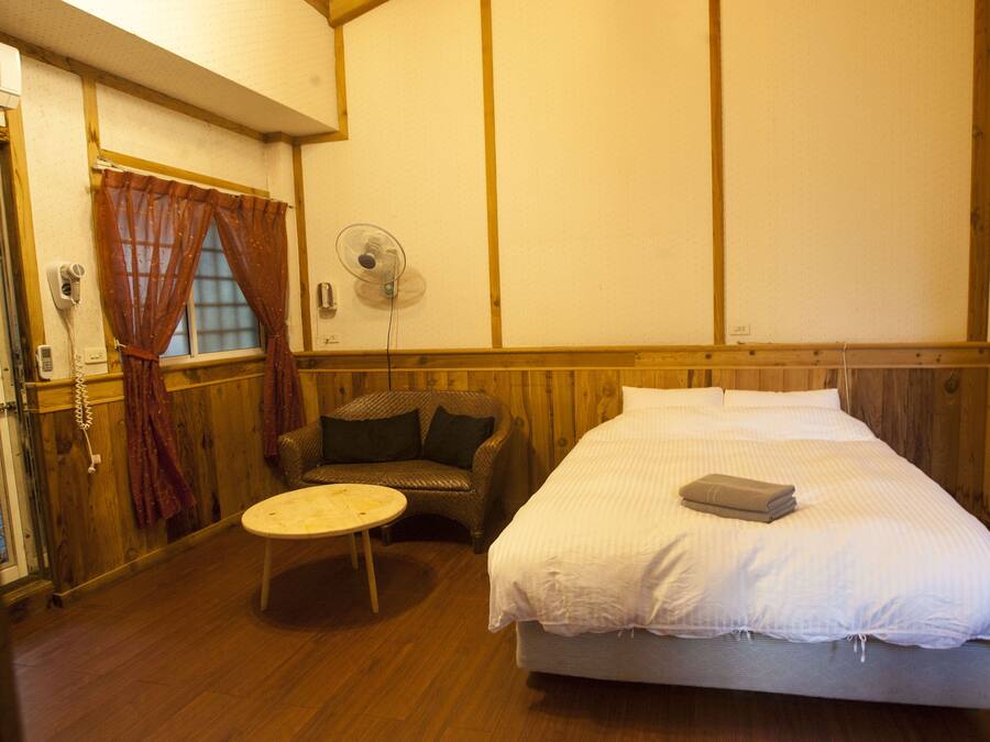 A room with a bed, a chair, a small table, a fan, and a window with curtains.