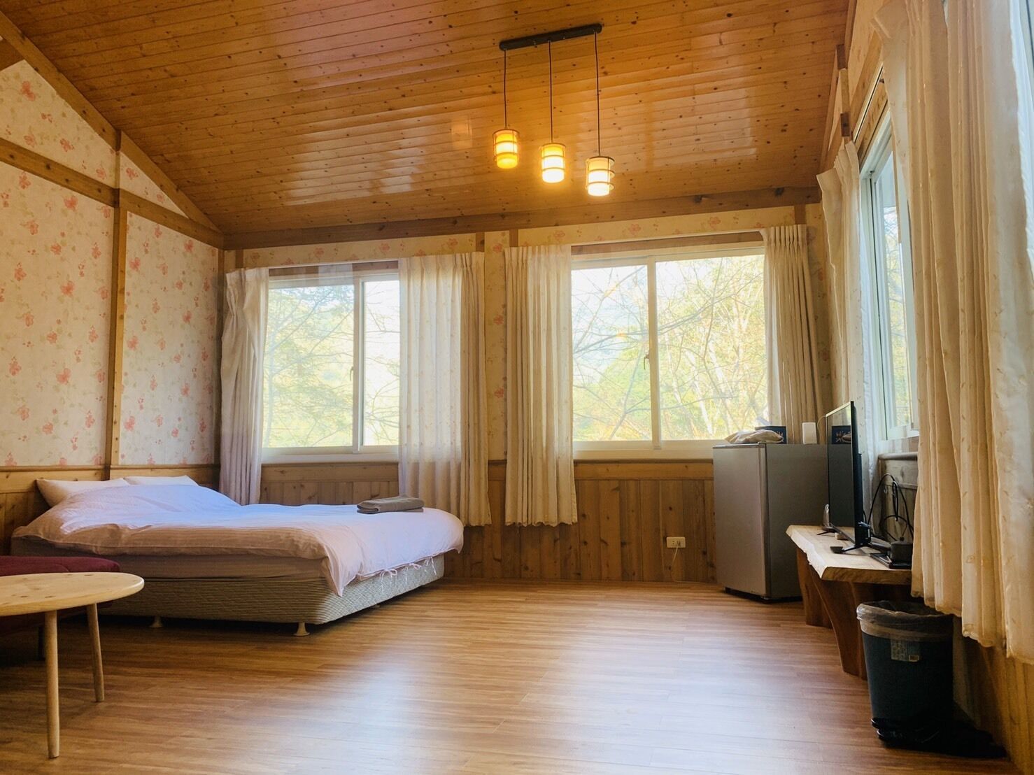 Une pièce confortable avec un lit, un bureau et un téléviseur. Elle comporte deux fenêtres avec des rideaux, et les murs et le plancher sont en bois.
