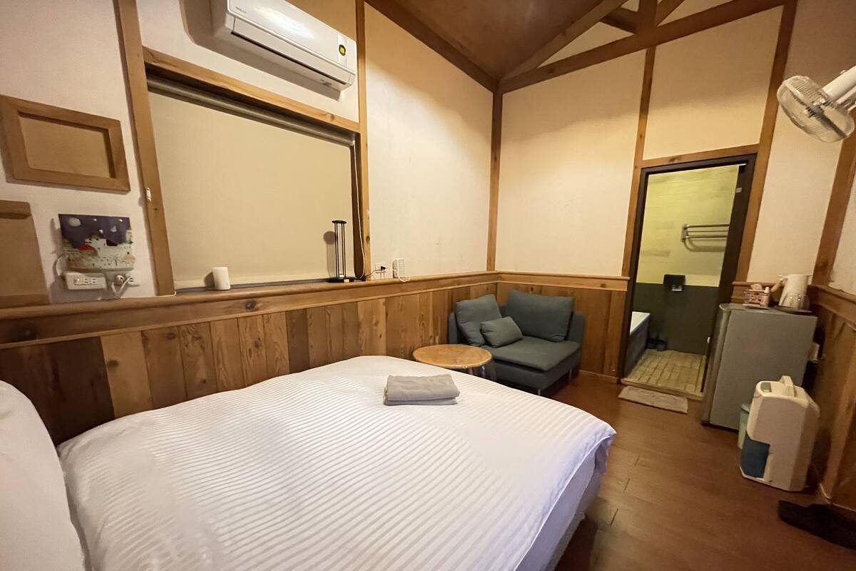 A hotel room with a bed, a small sofa, a round table, an air conditioning unit, a refrigerator, and a door leading to a bathroom.