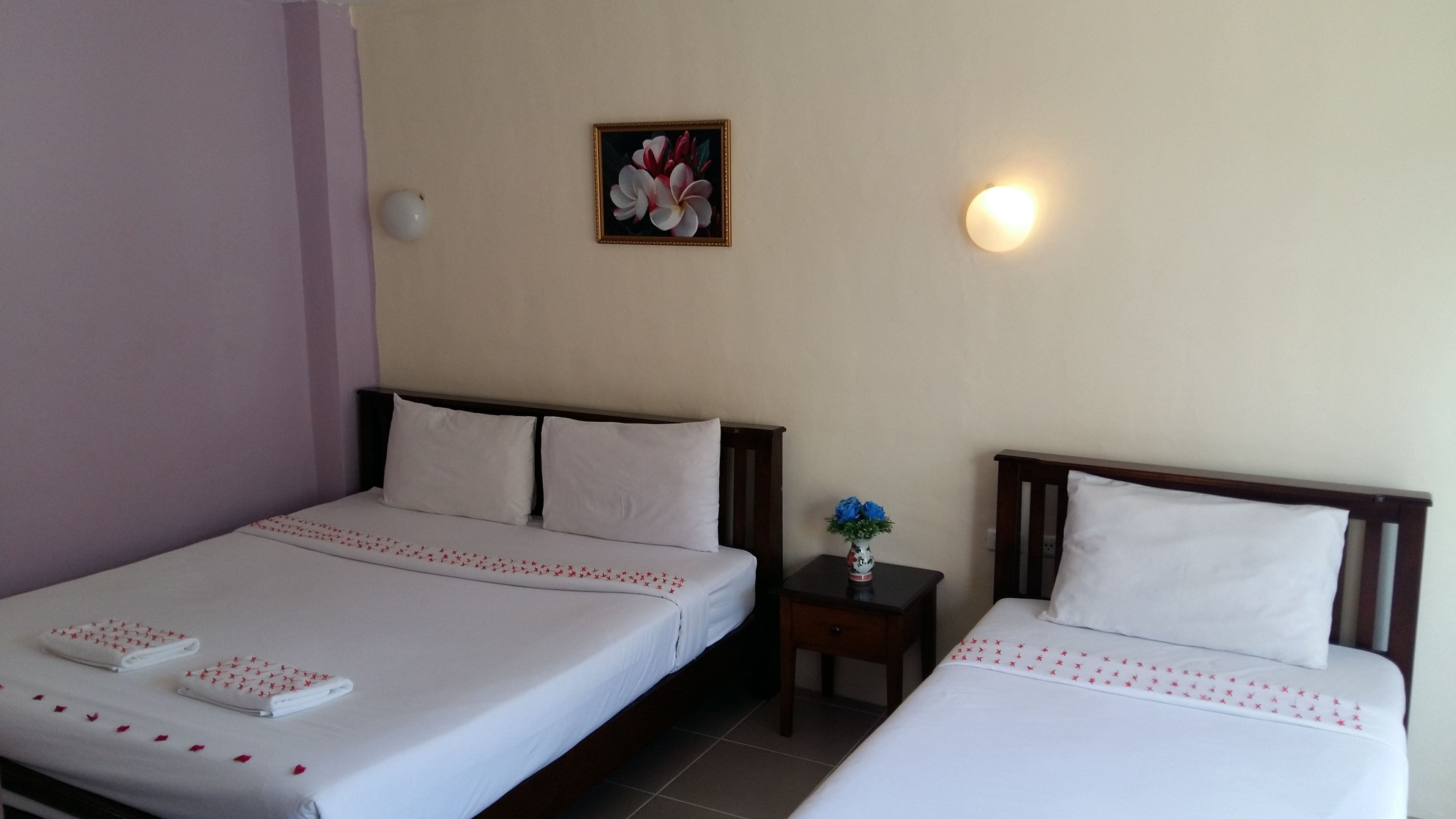 A hotel room with two single beds, a small bedside table with a vase of flowers, a wall-mounted light, and a framed painting above the beds.