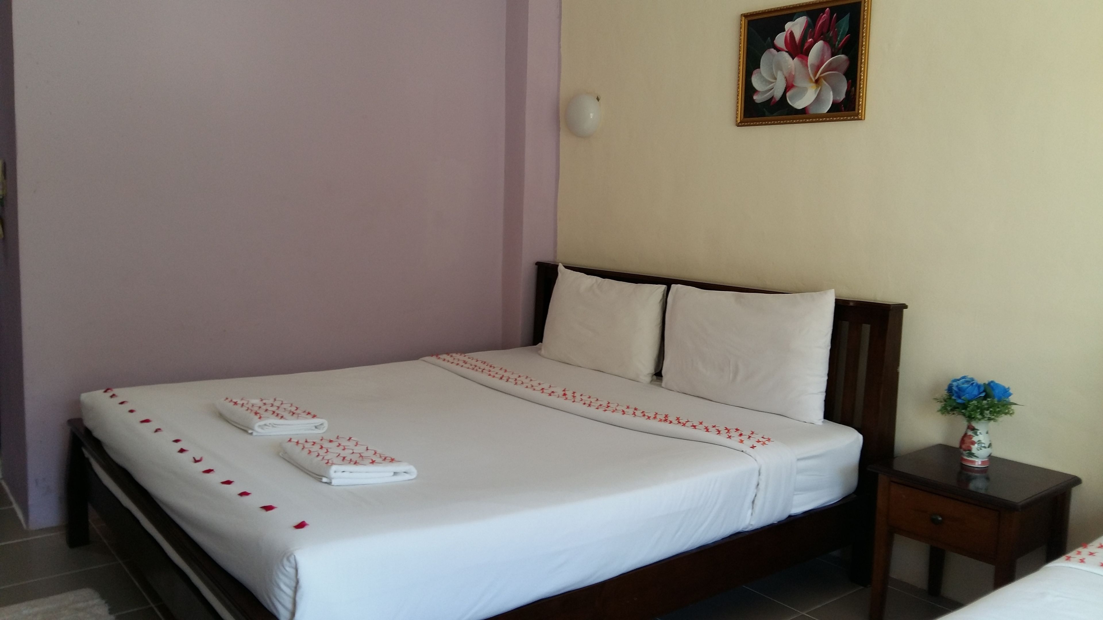 Una cama bien tendida con sábanas blancas y motivos florales rojos, un cabecero de madera y una mesita de noche con un jarrón de flores.