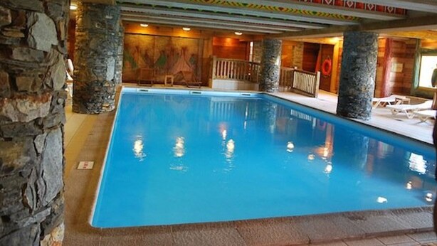 Una piscina interior con pilares de piedra, una terraza de madera y un diseño de techo colorido.