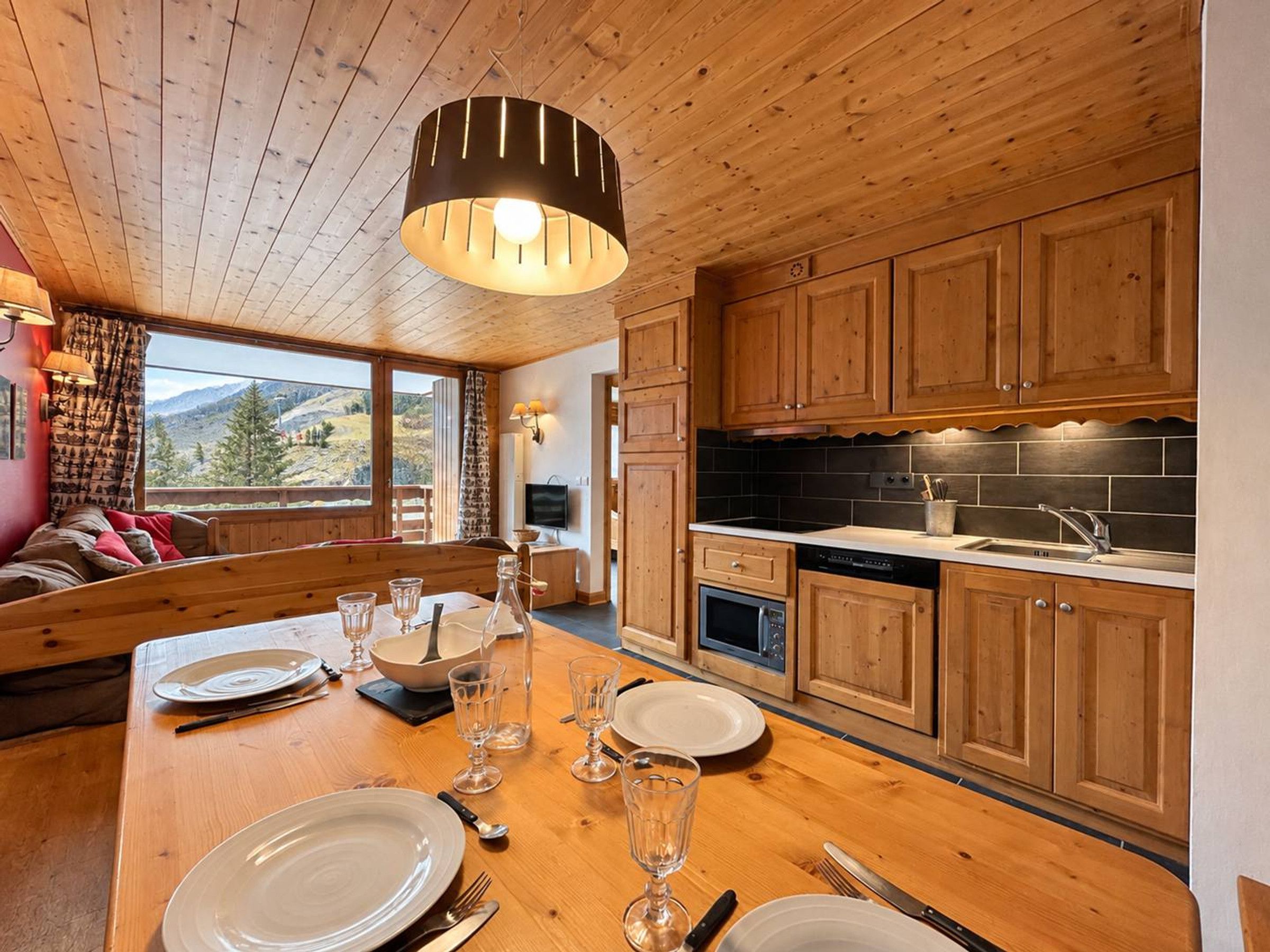 A wooden interior with a dining table set for four, a kitchen with wooden cabinets, and a view of the outdoors.