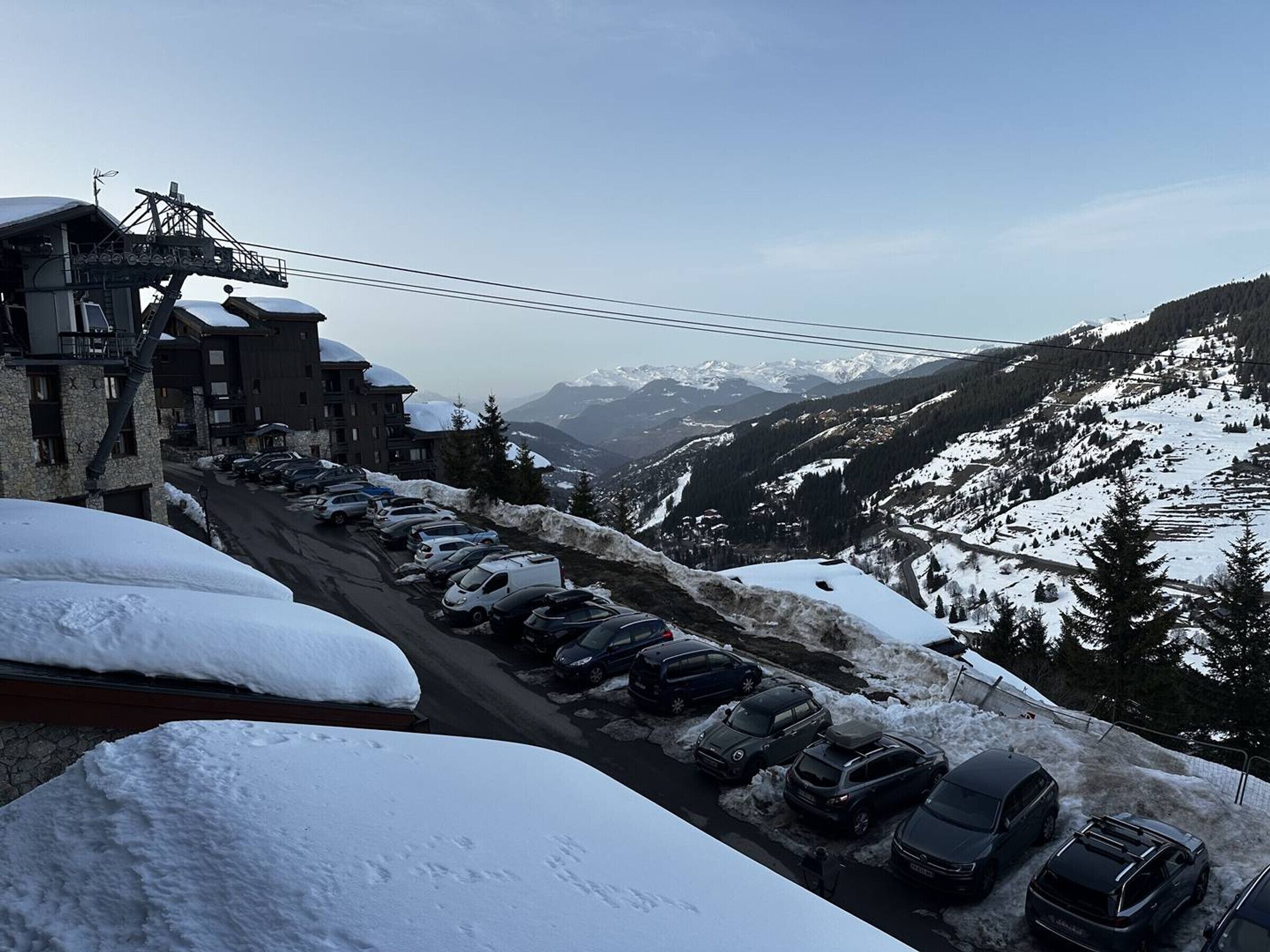Ein mit Schnee bedeckter Parkplatz mit Autos, ein Gebäude mit einer Seilbahn und eine bergige Landschaft im Hintergrund.