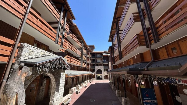 A narrow street flanked by multi-story buildings with balconies, stone walls, and a clear blue sky.