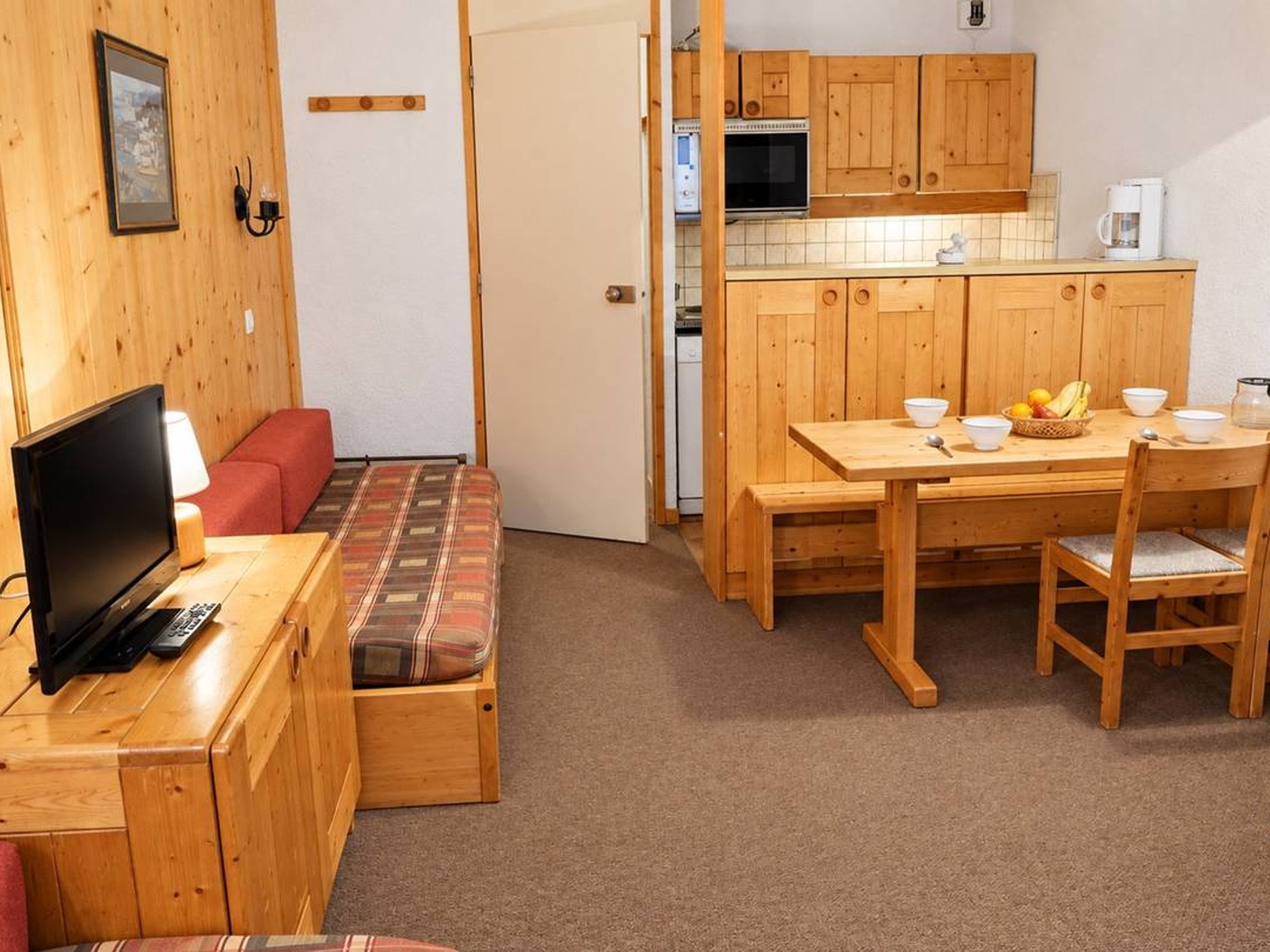 A compact room with a wooden bench, a television, a dining table with chairs, and a kitchen area with wooden cabinets and appliances.