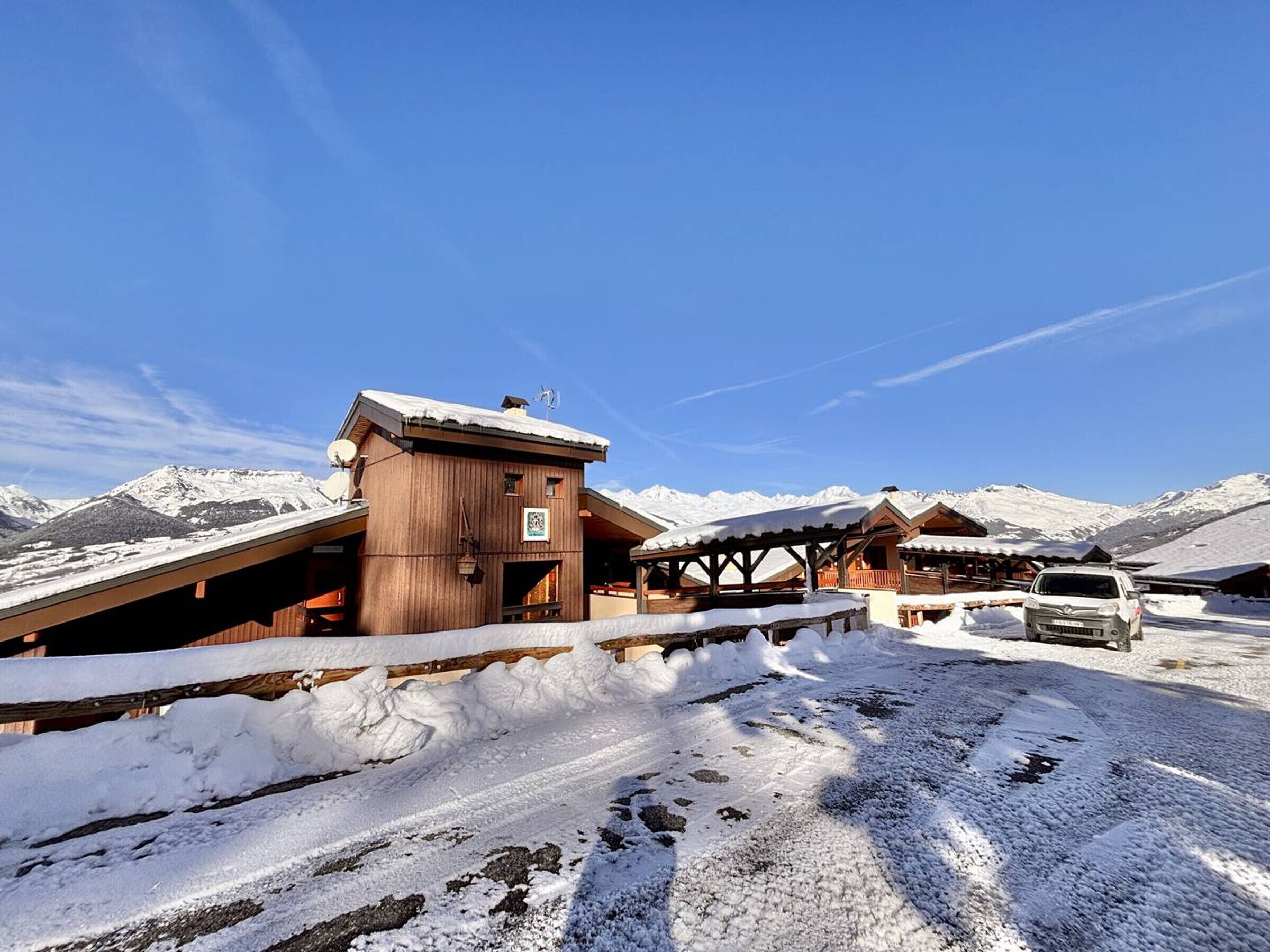 Un chalet en bois, dont le toit est recouvert de neige, avec une voiture garée devant et un paysage montagneux en arrière-plan.