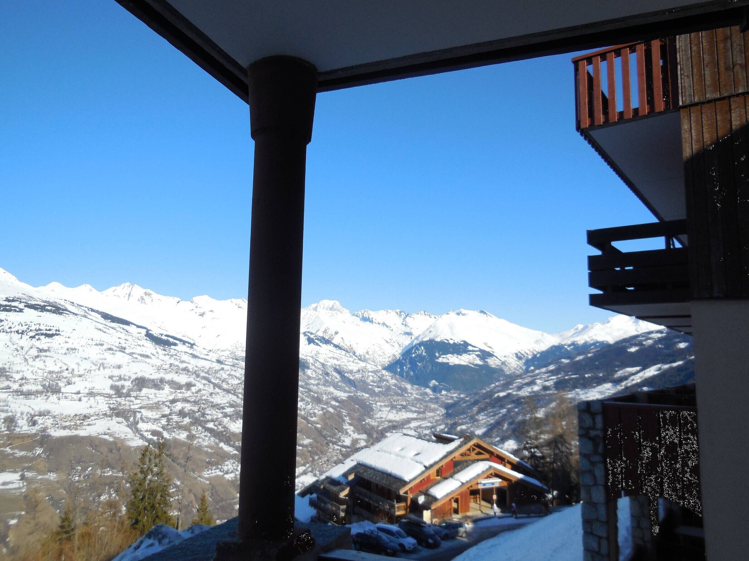 A mountain cabin with a snow-covered roof, a wooden balcony, and a view of snow-covered mountains.