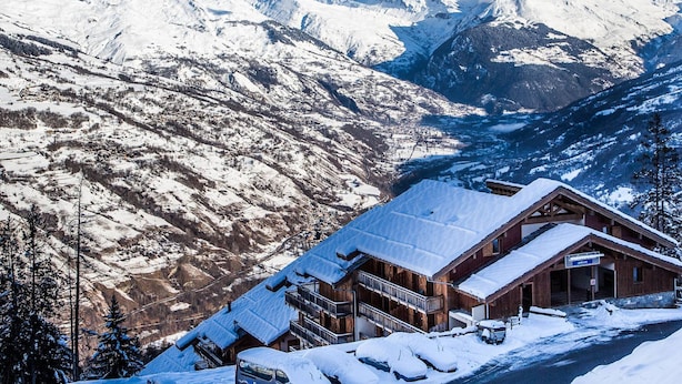 Un chalet en bois, dont le toit est recouvert de neige, situé au pied d’une chaîne de montagnes.