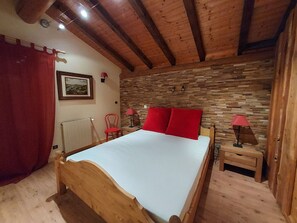 A bedroom with a wooden bed, a red headboard, a stone wall, a red curtain, a small table with a lamp, and a chair. - Comfortable chalet, mountain view - 9 guests (La Plagne Tarentaise)