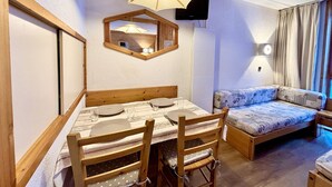 A cozy room with a wooden dining table set for four, a sofa with patterned cushions, a wall-mounted TV, and a ceiling light fixture.