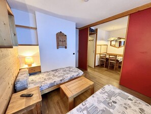 A compact room with a bed, a bench, a small table with a lamp, and a wooden wall-mounted shelf.
