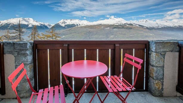 Un balcon avec une table et des chaises rouges, donnant sur une chaîne de montagnes enneigées.