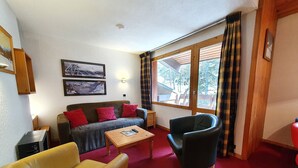 Una sala de estar acogedora con un sofá, un sillón y una mesa de centro. Hay dos cuadros en la pared, una ventana con cortinas y una lámpara en el techo.