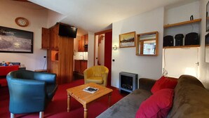 Una sala de estar con sofá, sillón y una pequeña mesa de madera. Hay un televisor montado en la pared y un cuadro encima.