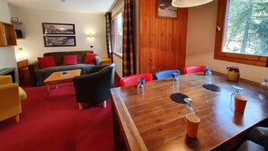 Una habitación acogedora con una mesa de madera, sillas rojas, un sofá con cojines rojos y un ventanal con vistas a árboles.