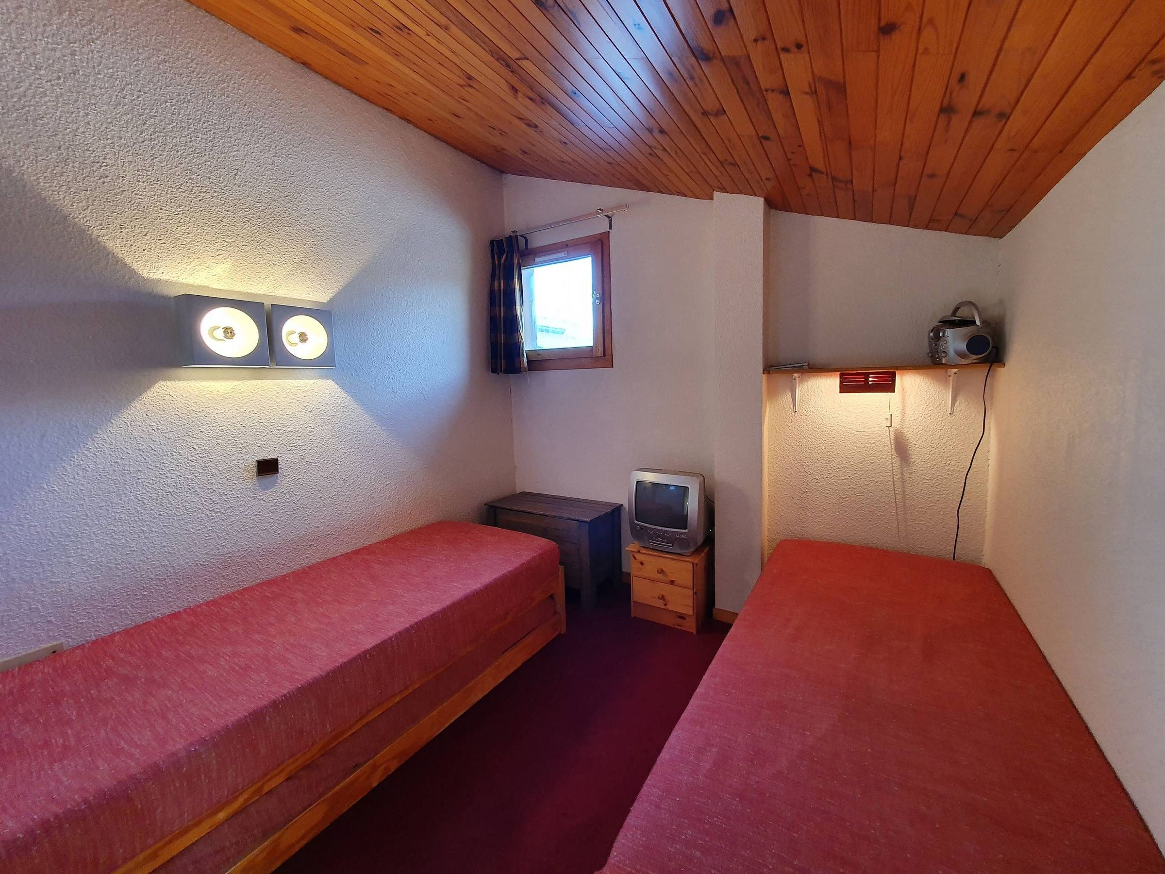 Una habitación pequeña y acogedora con dos camas individuales, techo de madera, un televisor pequeño sobre un mueble de madera y una ventana con cortinas.