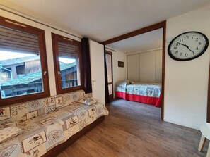A room with a wooden floor, a sofa with a patterned cover, a bed with a red bedspread, and a wall clock.