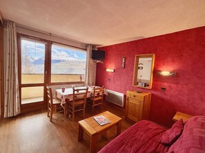 Um quarto com uma mesa e cadeiras de madeira, um pequeno armário de madeira, uma cama com um cobertor vermelho, uma televisão fixada na parede e uma janela com vista para montanhas.