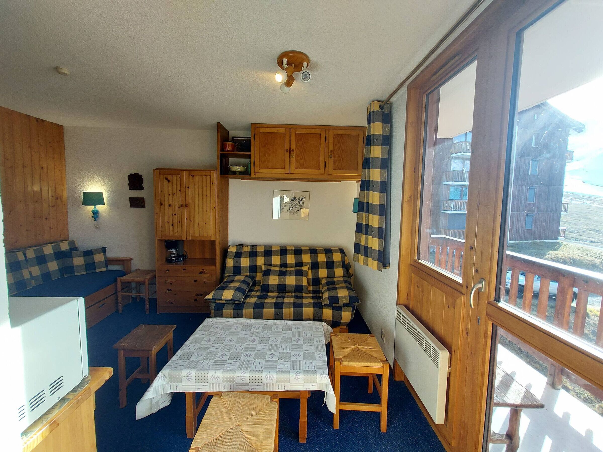 A cozy room with a bed, a table, a bench, a small table, a lamp, and a window with a view of buildings.