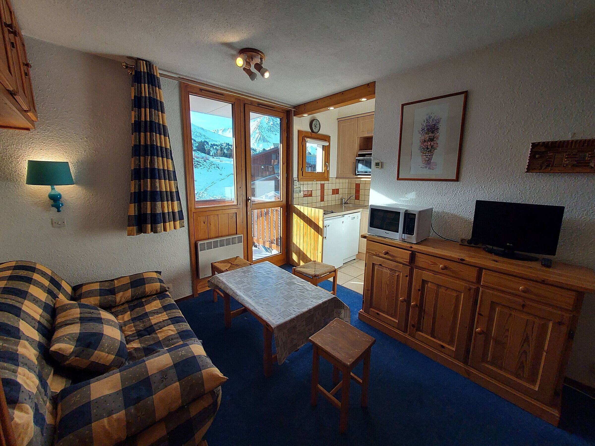 A cozy room with a sofa, a small table, a TV, and a kitchen area with a view of the mountains.