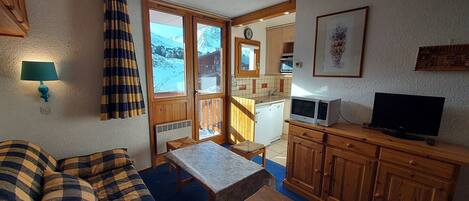 A cozy room with a sofa, a small table, a TV, and a kitchen area with a view of the mountains.