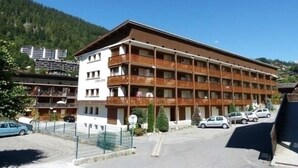 A multi-story hotel with balconies, a parking area, and a mountainous backdrop. - Cozy 3-Room Apartment, South Exposure, Village Center, Sleeps 6 (La Clusaz)