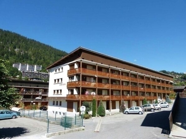 A multi-story hotel with balconies, a parking area, and a mountainous backdrop.