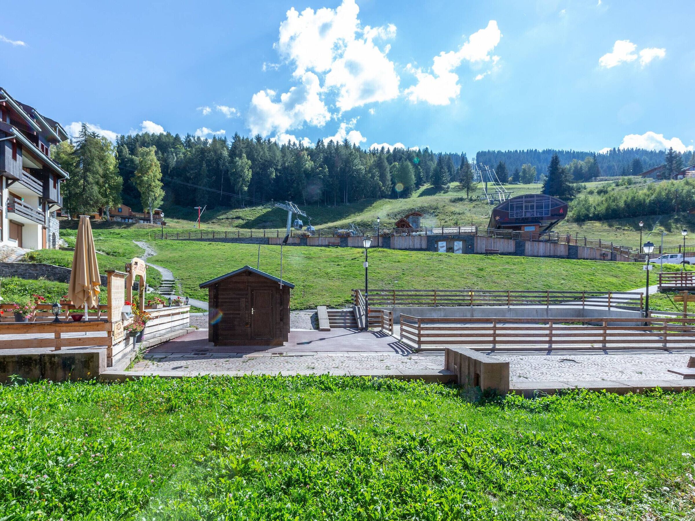 A wooden cabin with a metal roof, surrounded by a paved area with benches and a pathway leading to a ski lift.