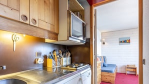 A compact kitchen with a sink, stove, and microwave. A wooden cabinet with a knife block is visible. A small dining area with a blue chair and a wooden table is seen in the background.