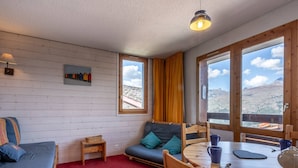 A cozy room with a wooden floor, a blue sofa, a small table with a blue mug, and a view of mountains through large windows.